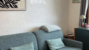 Un divano grigio con una coperta e due cuscini, un piccolo tavolino con una lampada, e un quadro appeso al muro.