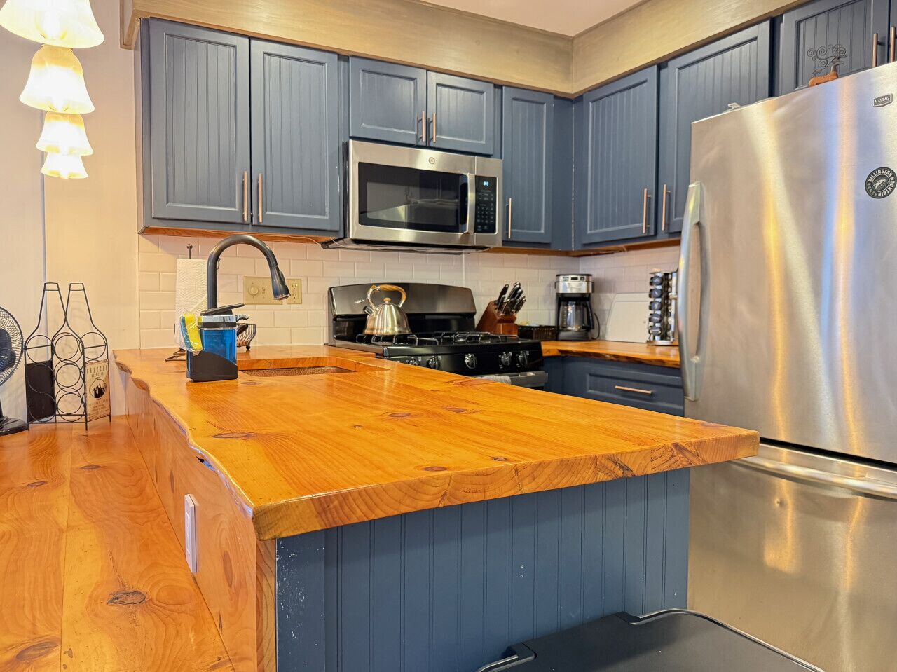 A kitchen with blue cabinets, a stainless steel refrigerator, a microwave, and a wooden countertop.