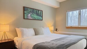 Ein Schlafzimmer mit einem großen Bett, einem Nachttisch, einer Lampe, einem Fenster mit Blick auf Bäume und einem Bild an der Wand.