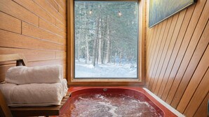 A wooden cabin interior with a large window, a red hot tub, and a bench with a towel. - Mountain Moose Chalet- indoor hot tub, FirePit (Killington)