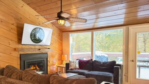 A cozy cabin interior with a fireplace, a ceiling fan, and a large window. - Mountain Moose Chalet- indoor hot tub, FirePit (Killington)