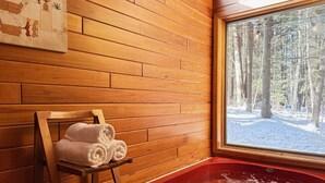 A wooden cabin interior with a wooden chair holding towels, a red bathtub, and a window with a snowy forest view. - Mountain Moose Chalet- indoor hot tub, FirePit (Killington)