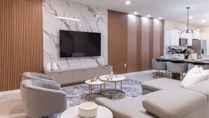 Ein modernes Wohnzimmer mit Flachbildfernseher, grauem Polsofa, runden Couchtischen und einer Küchenzeile mit weißen Schränken und einer Mikrowelle.