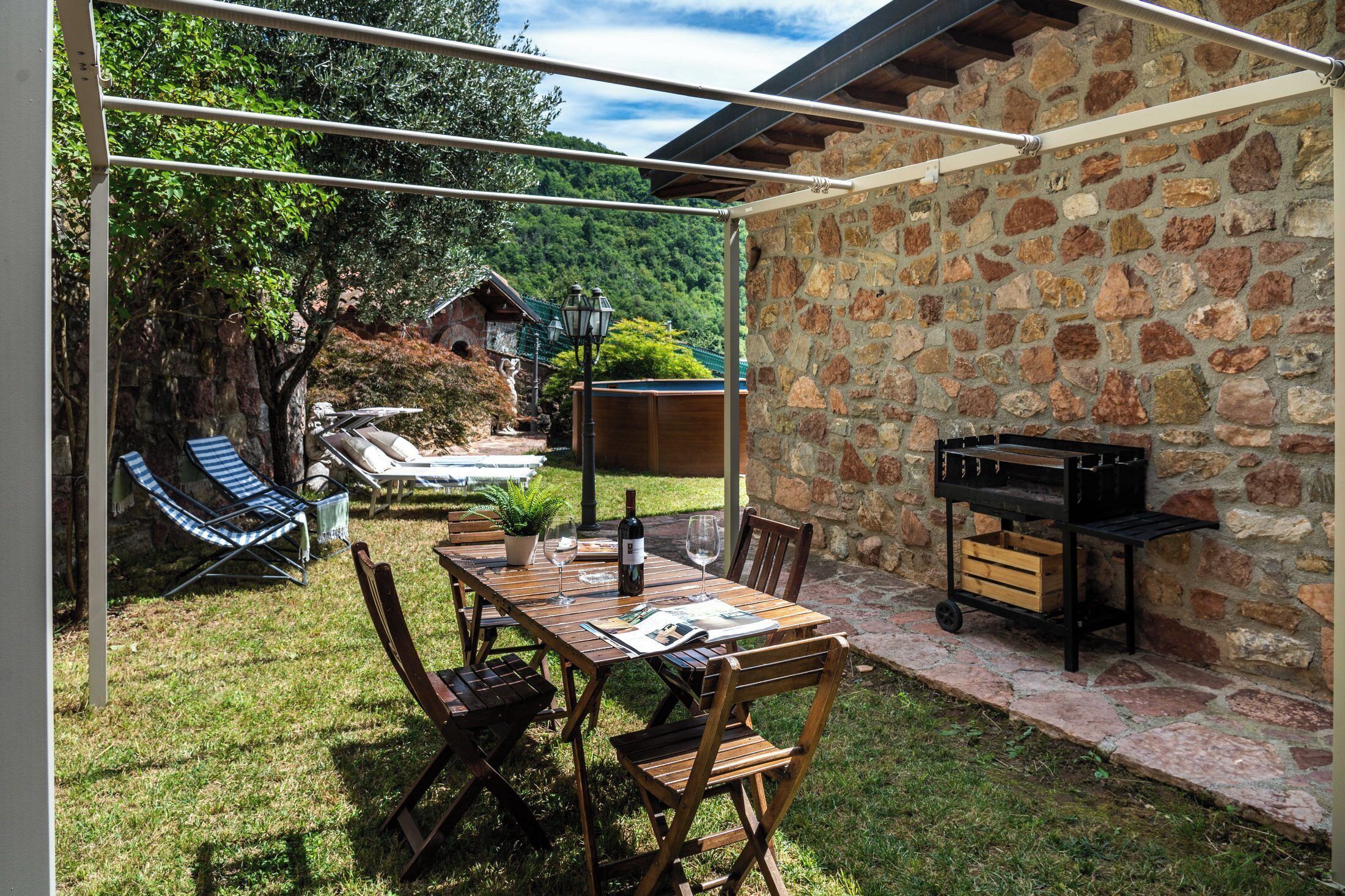 An outdoor patio with a stone wall, a wooden table with chairs, a grill, and a hammock.