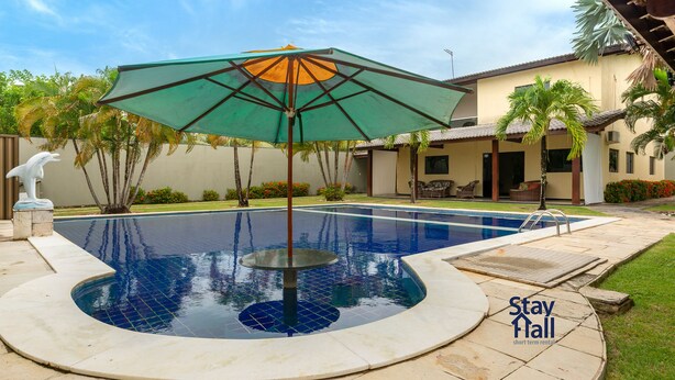 Ein Swimmingpool mit einem runden Whirlpool, umgeben von einer Terrassenanlage mit einem grünen Sonnenschirm. Im Hintergrund sind eine Delphin-Statue, Palmen und ein Gebäude mit einem Ziegeldach zu sehen.