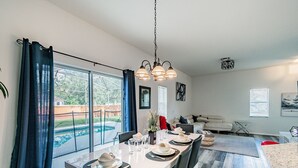 Ein Essbereich mit einem großen Tisch, gedeckt für vier Personen, einer Hängeleuchte und Blick auf einen Swimmingpool durch große Fenster.