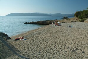 Una platja sorrenca amb gent prenent el sol i banyant-se, una costa rocosa i muntanyes a la distància.