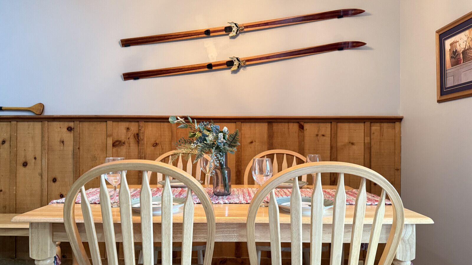 A wooden dining table with four chairs, a vase with flowers, and two skis mounted on the wall above.