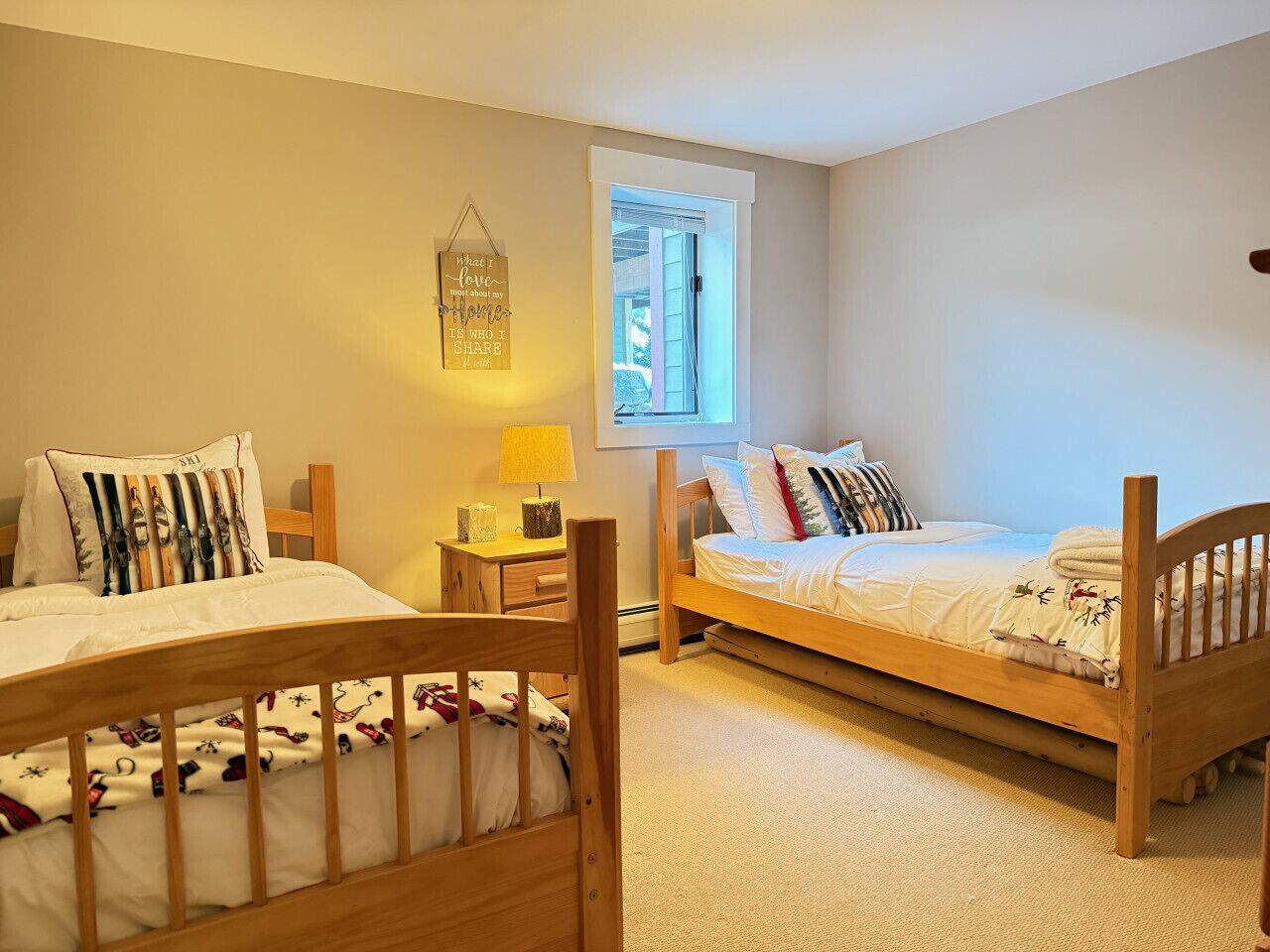 A bedroom with two wooden beds, a bedside table with a lamp, a window with blinds, and a wall-mounted light fixture.
