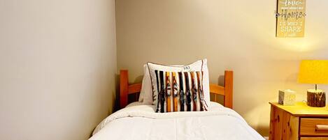 A neatly made bed with a white duvet and patterned bedding, a wooden headboard, and a bedside table with a lamp and a small decorative item.