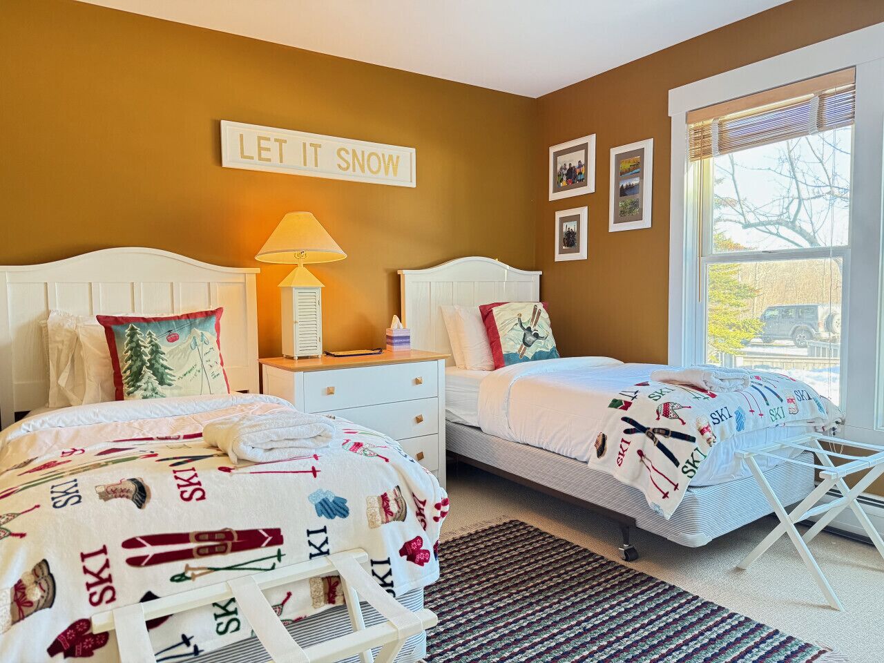 A bedroom with two beds, a window with blinds, a bedside table with a lamp, and a wall sign that reads "LET IT SNOW".