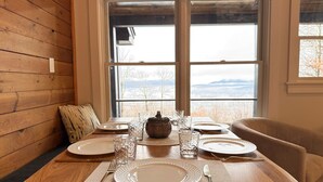 A dining table set with plates, glasses, and cutlery, positioned in front of a window with a view of trees and mountains. - Sunset Notch- Hot tub, Cold Plunge, Hiking, Views! (Mendon)