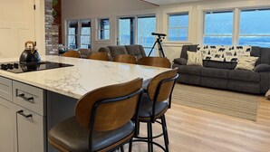 A modern kitchen with a marble countertop and black kettle, a dining area with wooden chairs, and a gray sofa with a bear-themed throw pillow. - Sunset Notch- Hot tub, Cold Plunge, Hiking, Views! (Mendon)