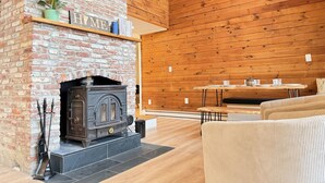 A cozy living room with a brick fireplace, wooden walls, a dining table, and a comfortable sofa. - Sunset Notch- Hot tub, Cold Plunge, Hiking, Views! (Mendon)