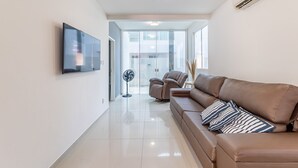 Una sala de estar moderna con un televisor de pantalla plana grande, un sofá de cuero marrón con almohadones a rayas, un ventilador de pie y un cerramiento de vidrio que da a un espacio luminoso.