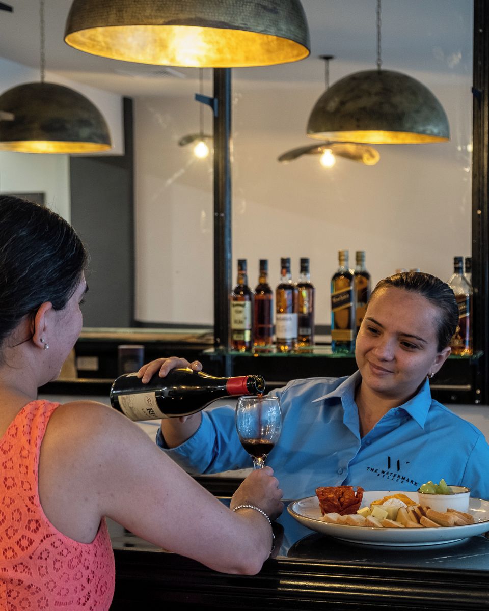 A person pouring wine into a glass at a bar with bottles in the background.