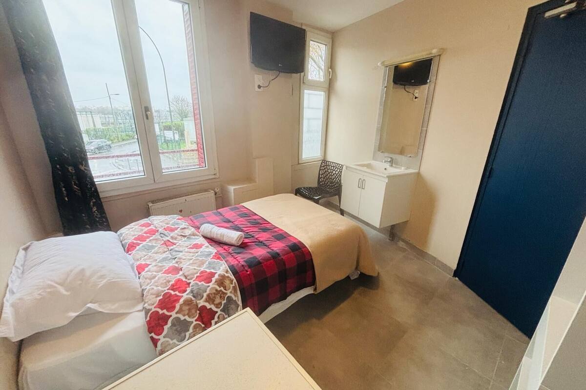 A bedroom with a bed, a bedside table, a window, a TV, a sink, and a mirror.