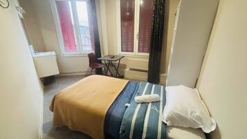 A small, neatly arranged bedroom with a bed, a chair, a table, and a window with shutters.