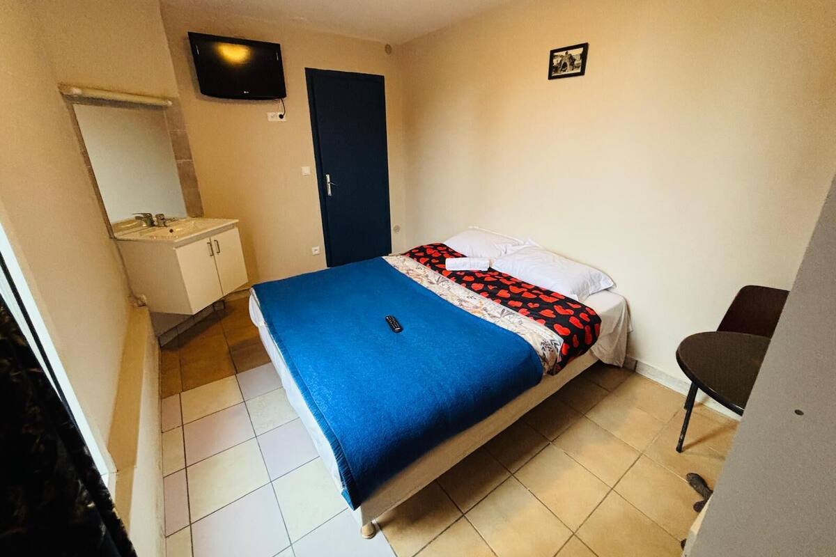 A bedroom with a bed, a bedside table, a chair, a TV, and a bathroom with a sink and mirror.