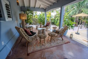 A covered outdoor seating area with wicker furniture, a lamp, and a view of tropical plants. - Little Bali Curaçao: Exotic Caribbean Oasis (Grote Berg)
