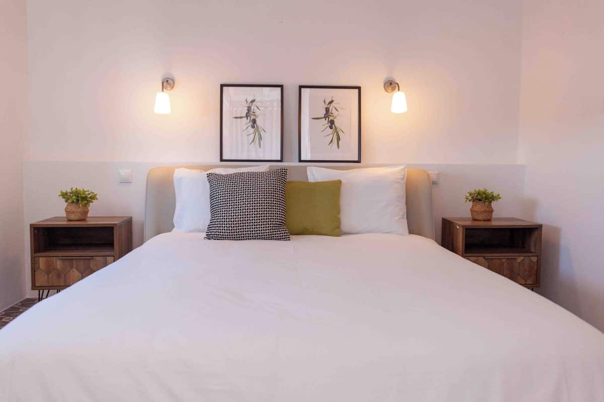 A neatly made bed with white bedding, two framed pictures on the wall, and two bedside tables with potted plants.