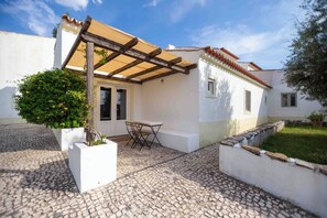 A white building with a tiled roof, a pergola, a table with chairs, and a cobblestone patio.