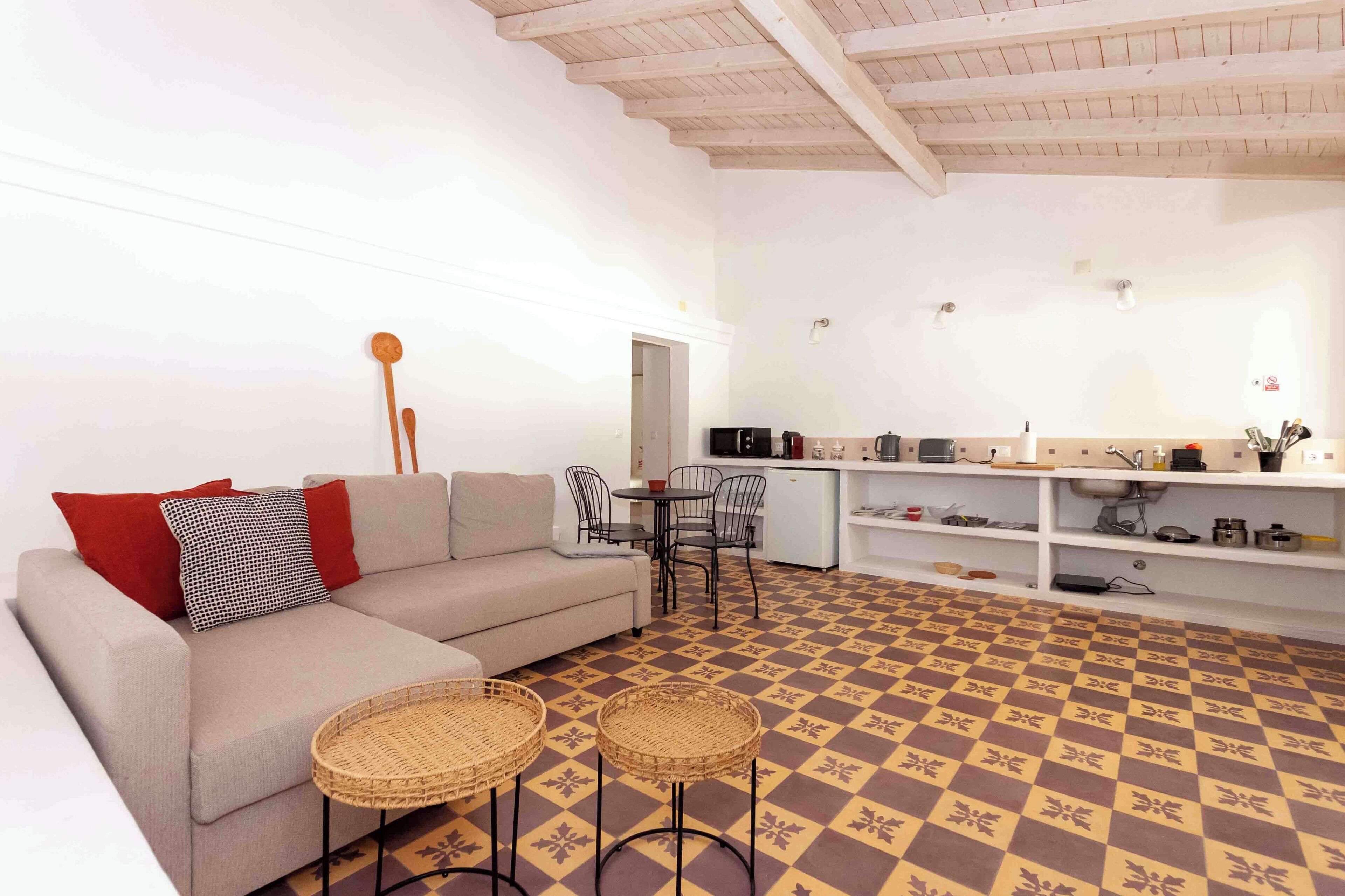 Una sala de estar moderna con un sofá, dos mesas de mimbre y una zona de cocina con mostrador y electrodomésticos.