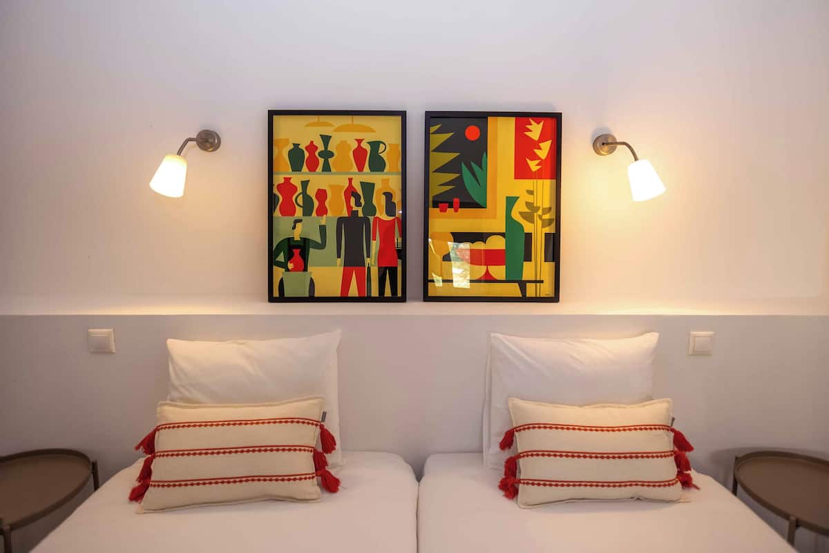 A hotel room with two beds, each with white bedding and red tassel pillows. There are two wall-mounted artworks above the beds, and two wall-mounted lamps on either side.