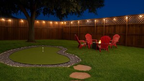 Un jardin avec un terrain d’entraînement au golf, des chaises rouges, un foyer et une clôture en bois.