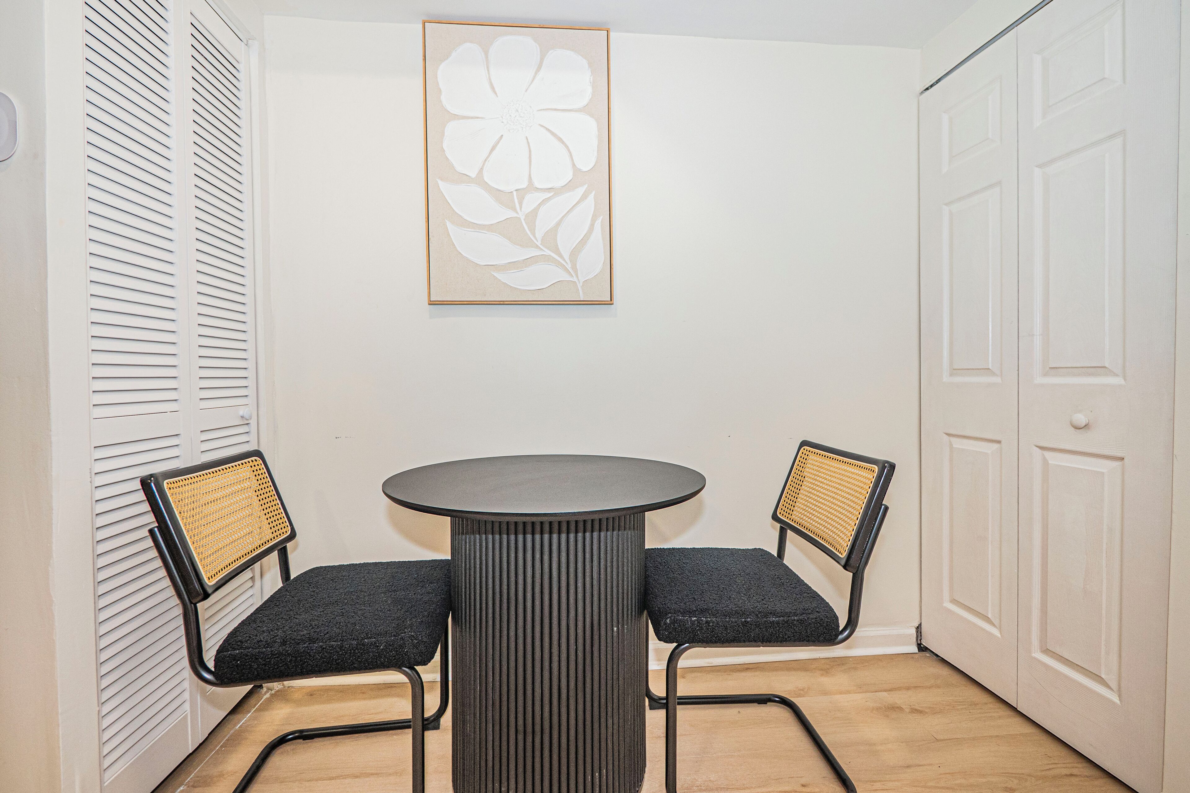 Ein Zimmer mit einem runden Tisch, zwei Stühlen, einem gerahmten Blumenbild an der Wand und einer weißen Tür.