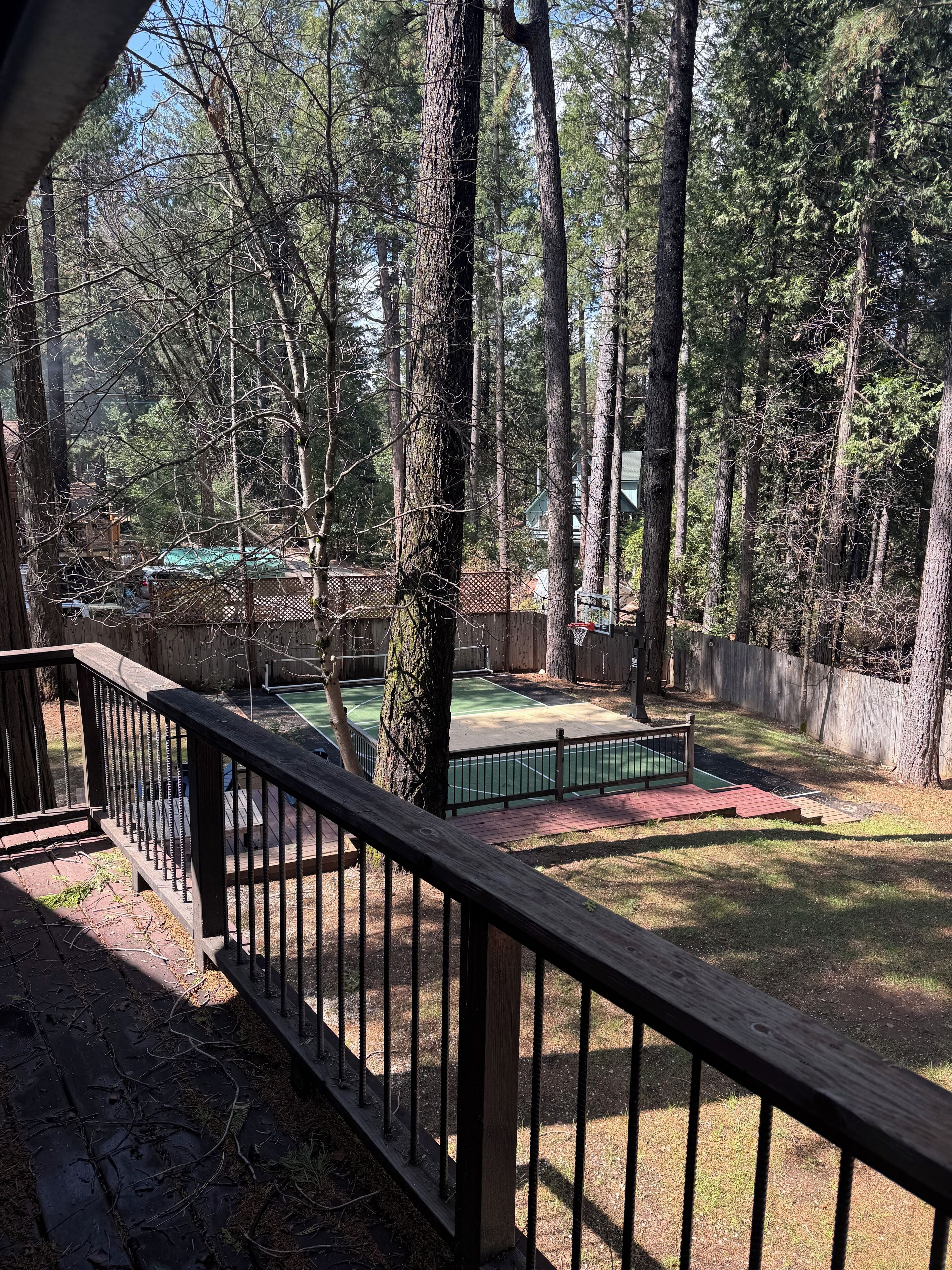 A wooden deck with a metal railing overlooking a backyard with a green tennis court and a fenced play area.
