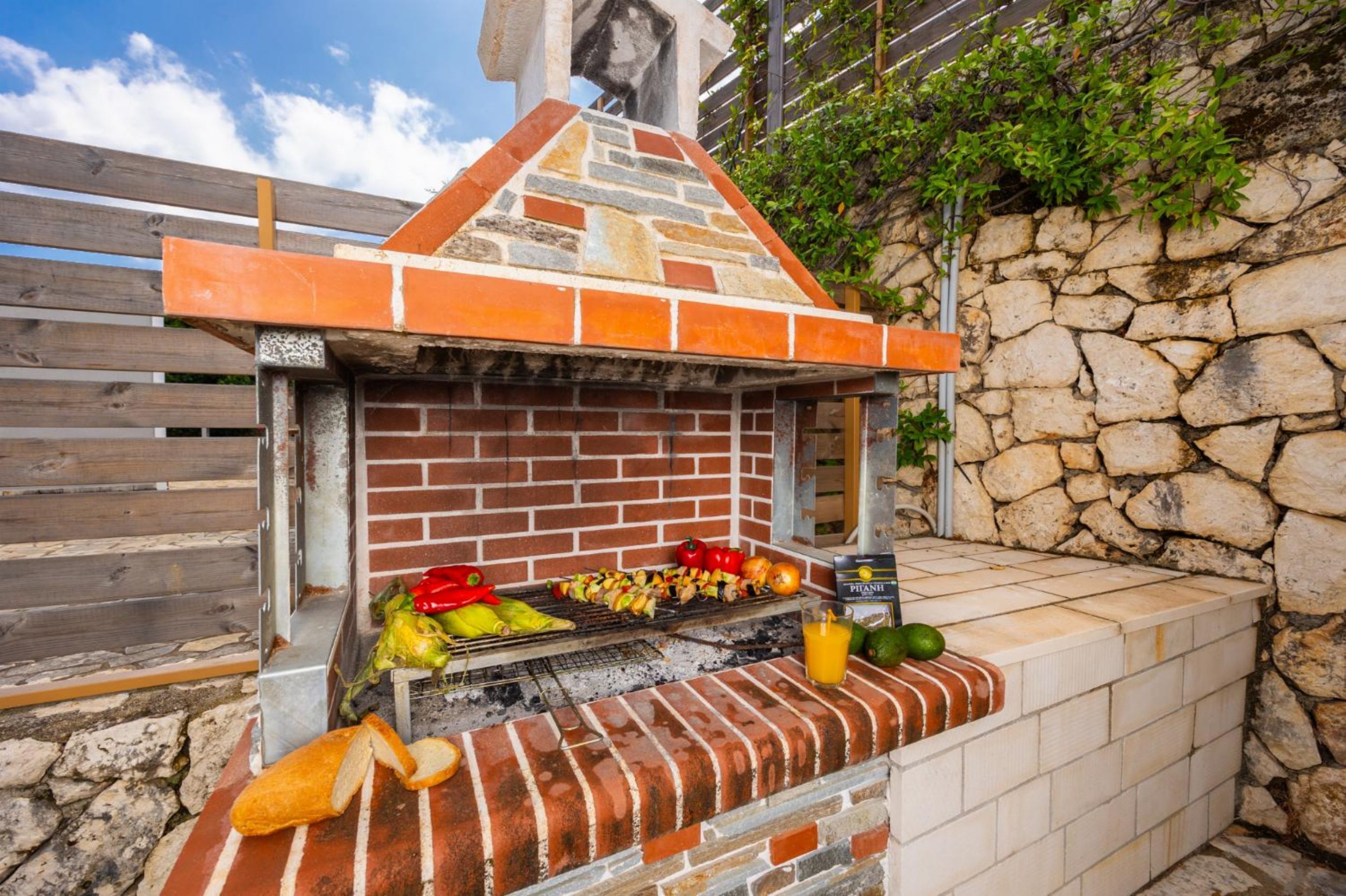 Ein Grill aus Ziegelsteinen im Freien mit Gemüse und Spießen, eine Steinmauer und ein Holzzaun.