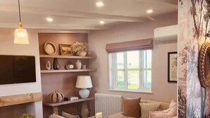 A cozy living room with a sofa, a coffee table, and a TV mounted on the wall. There is a window with a view of the outdoors, and a fireplace with a mantel. - The Fuller's Shed All Weather Couples Rural Retreat with Private Hot tub (Dadlington)