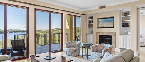 A spacious living room with a large sliding glass door leading to a balcony with a view of the water. There is a fireplace, a television mounted on the wall, and a bookshelf.