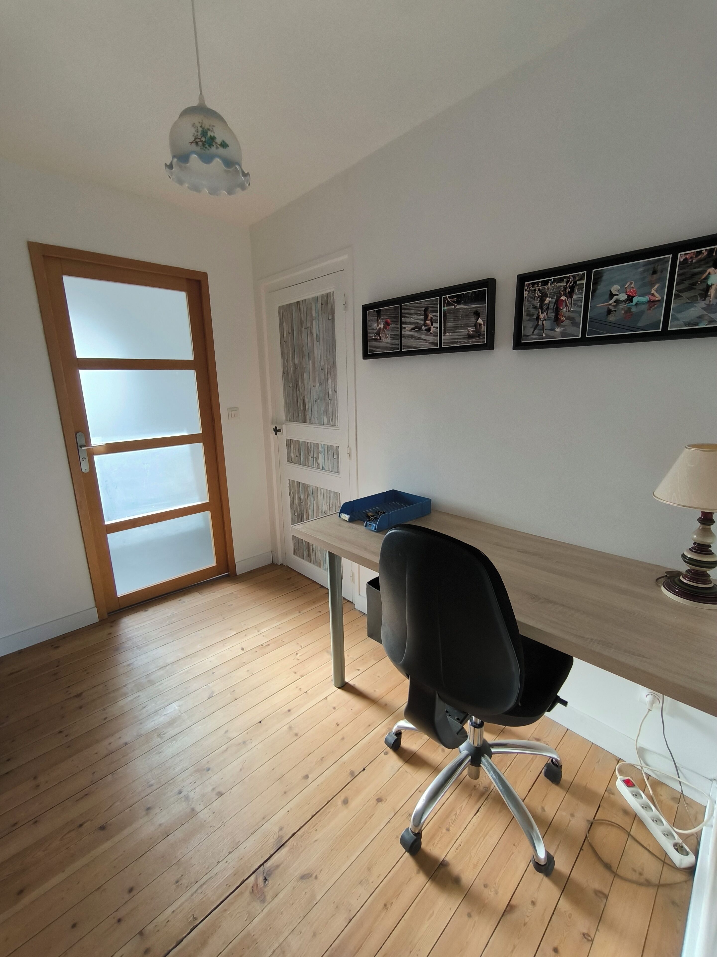 A room with a wooden floor, a desk with a chair, a wall-mounted shelf, a door with frosted glass panels, and a ceiling light fixture.