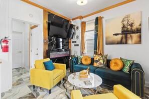 A living room with a yellow armchair, a blue sofa with decorative pillows, a flat-screen TV, a fireplace, and a painting on the wall. - VIP LUX-2 Bedroom Apart-15 min NYC-Shared Parking (Fairview)