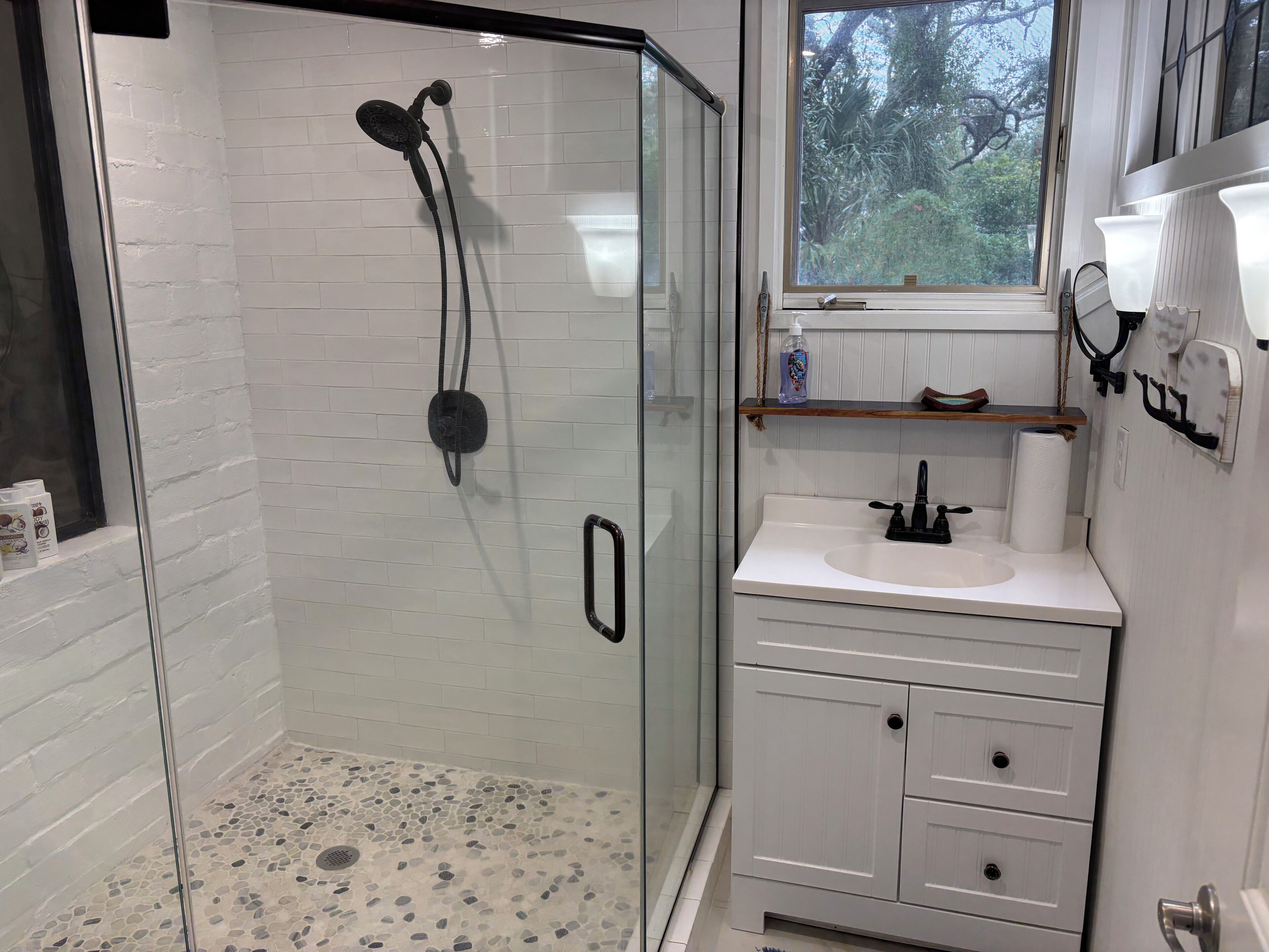 Une salle de bain moderne avec une douche à parois de verre, des murs recouverts de carreaux blancs, un lavabo blanc et une fenêtre donnant sur la verdure.