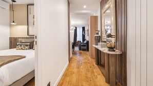 Un couloir d’hôtel avec un plancher en bois, un lit doté d’une tête de lit blanche, un grand miroir et une petite table ornée d’objets décoratifs.