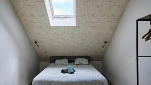 A small, narrow room with a sloped ceiling and a window. There is a bed with a patterned bedspread and pillows, a bench, and a wardrobe with hanging clothes. - En déplacement 10, les combles wifi gratuit (Tourcoing)