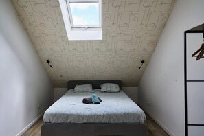 A small, narrow room with a sloped ceiling and a window. There is a bed with a patterned bedspread and pillows, a bench, and a wardrobe with hanging clothes. - En déplacement 10, les combles wifi gratuit (Tourcoing)