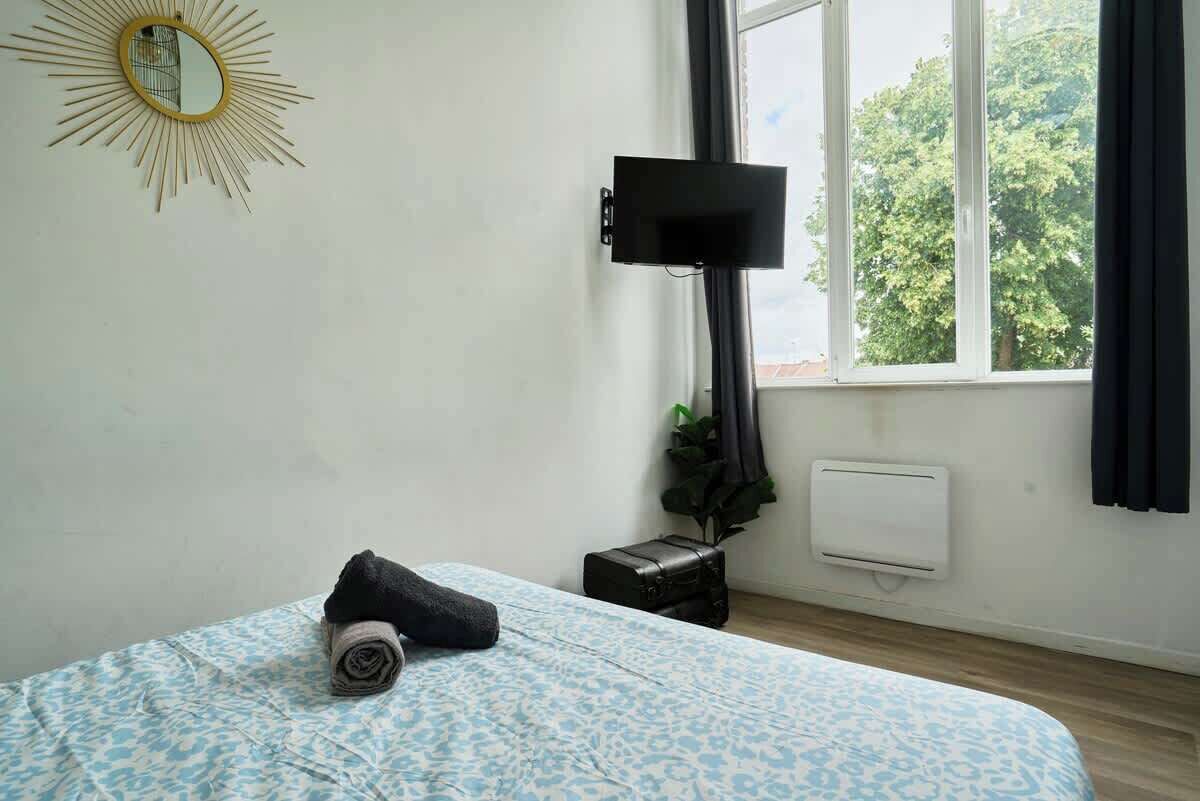 A bedroom with a bed, a wall-mounted TV, a window with curtains, a radiator, and a small plant.