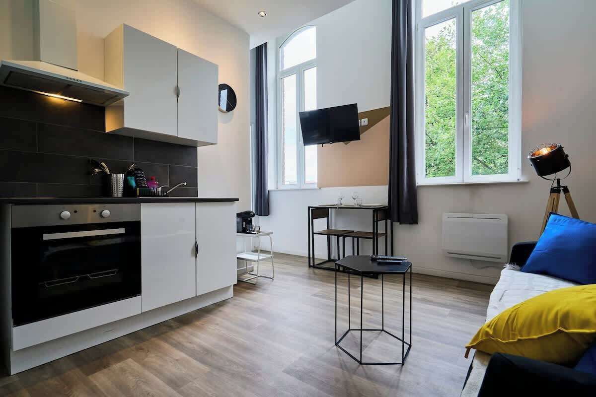 A modern kitchen with a built-in oven, a dining area with a table and chairs, a bed with a yellow pillow, and a TV mounted on the wall.