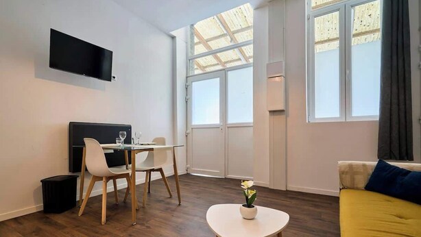Un salon moderne avec un téléviseur à écran plat, un coin repas comprenant une table et des chaises, une petite table blanche ornée d’un vase, et un canapé avec un coussin.
