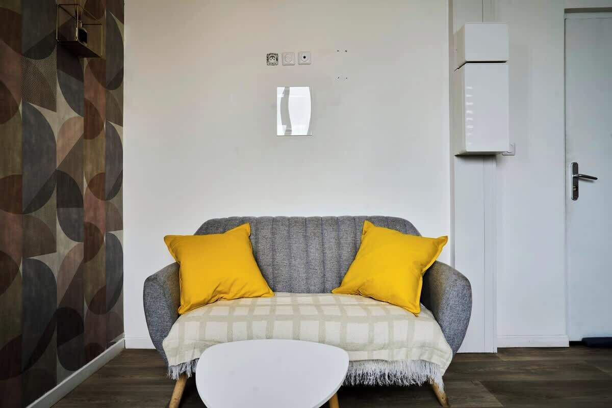 Een woonkamer met een grijze bank, twee gele kussens, een witte salontafel en een behang met patroon.