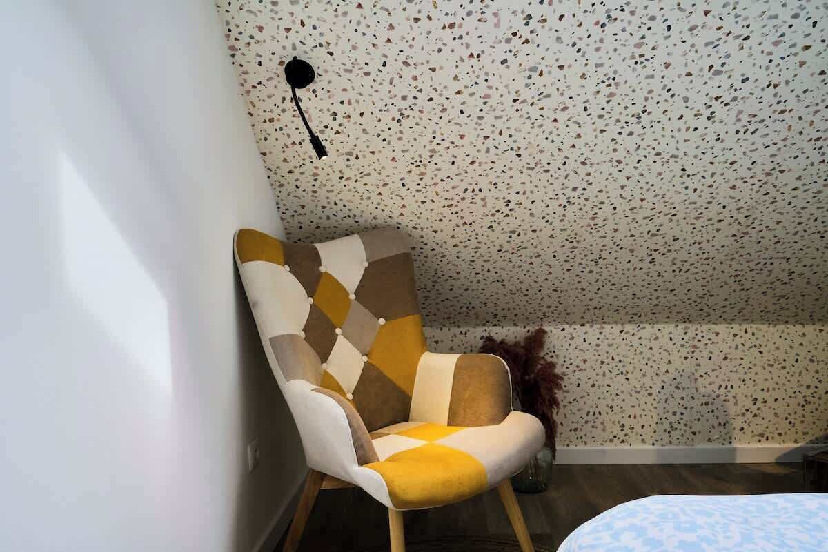 A corner of a room with a patterned ceiling, a chair with a geometric upholstery design, and a wall-mounted light fixture.