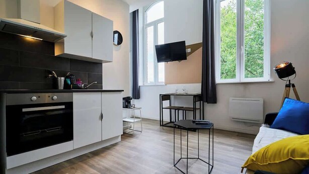 A modern kitchen with a built-in oven, a dining area with a table and chairs, a bed with a yellow pillow, and a TV mounted on the wall. - Modern Apt Near Tourcoing Sta. w/ Fast WiFi (Tourcoing)