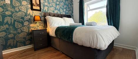 A bedroom with a bed, a nightstand, a lamp, a framed picture, and a window with curtains.