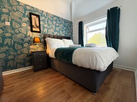 Un dormitorio con cama, mesita de noche, lámpara, un cuadro enmarcado y una ventana con cortinas.