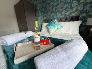 Una cama bien tendida con una bandeja que contiene una botella de vino, copas y una caja roja.