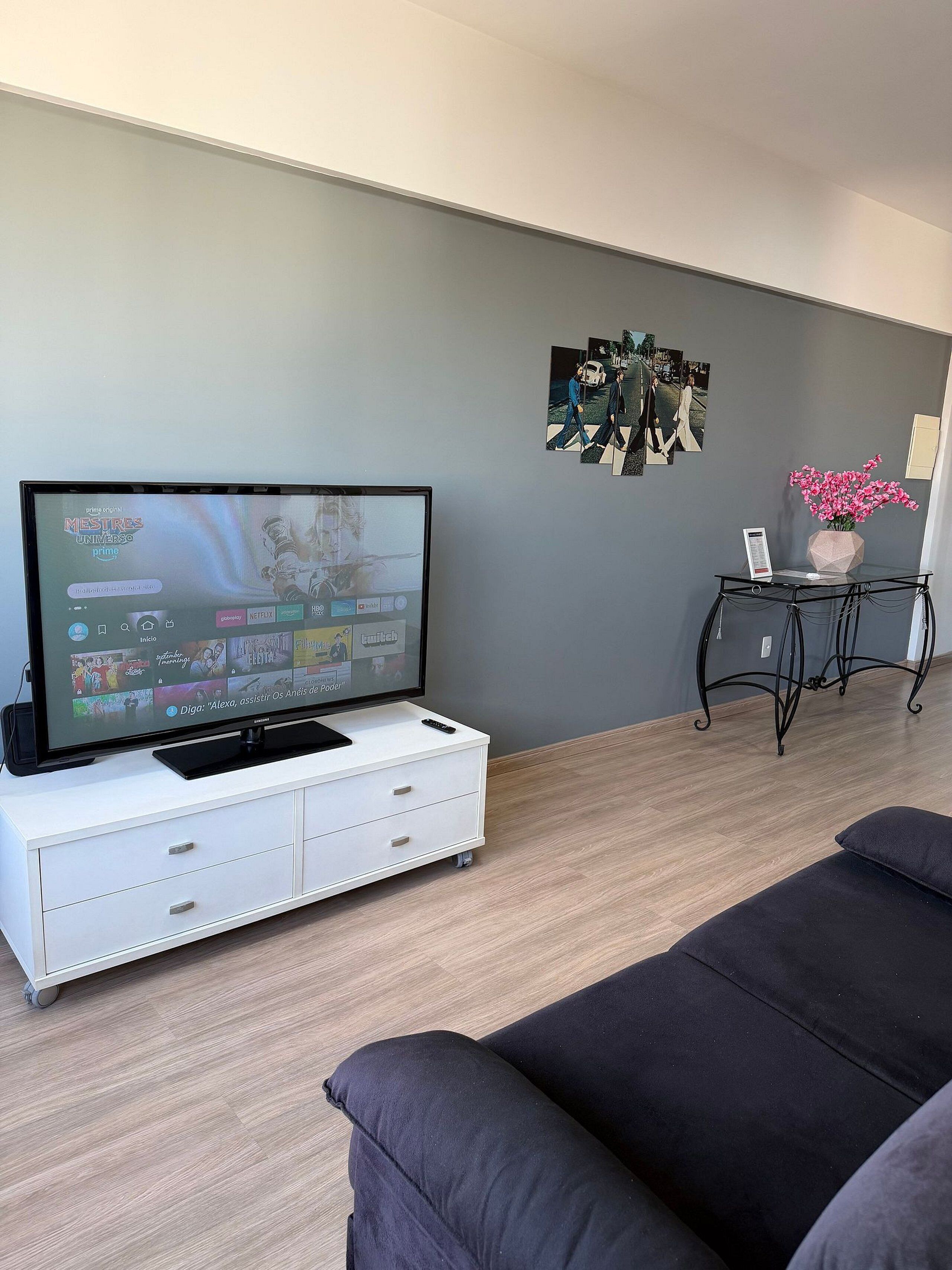 Un salon avec un téléviseur à écran plat posé sur un meuble blanc, un canapé noir, une petite table sur laquelle se trouve un vase de fleurs et une œuvre d’art accrochée au mur.